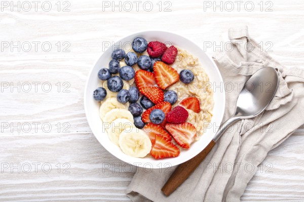 Oatmeal porridge with fruit and berries in bowl with spoon on white wooden background table top view, homemade healthy breakfast cereal with strawberry, banana, blueberry, raspberry. Space for text, food photography