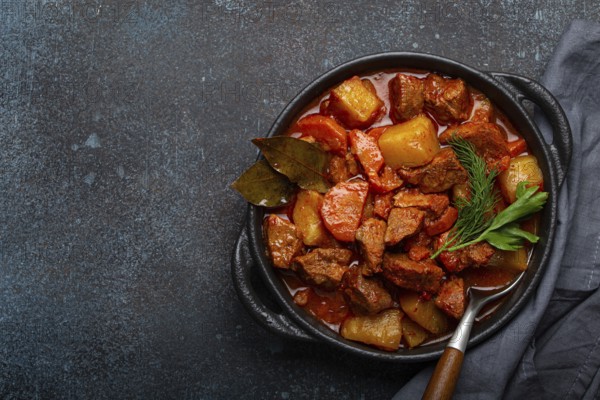 Beef meat stew with potatoes, carrot and delicious gravy in black casserole pot with bay leaves and fresh green herbs with spoon on dark rustic concrete background from above, space for text, food photography