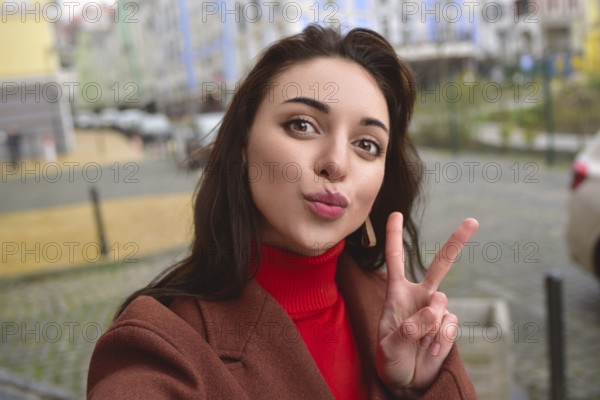 Charming smiling lady in a brown autumn coat takes a selfie on the street with a peace sign
