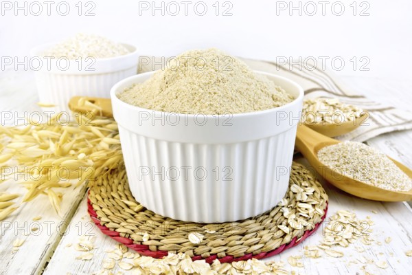 Oat flour in white bowl, oatmeal and bran in a spoon, stalks of oats, napkin against the background of wooden boards