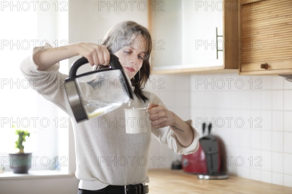 Woman pouring water into a cup in a bright kitchen with kettle, energy-saving, environmentally friendly