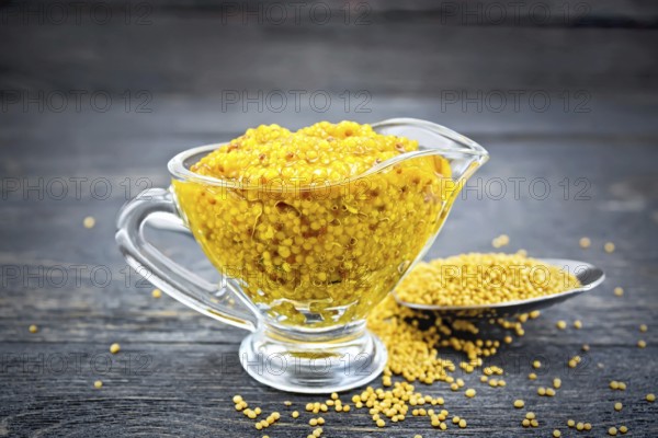 Mustard Dijon sauce in a glass sauceboat and mustard seeds in a spoon on a black wooden board background