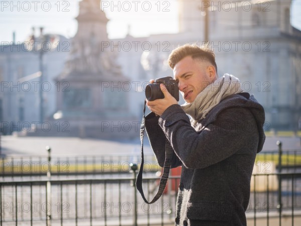 Handsome young male photographer taking photograph with professional photo camera hanging from his neck, outdoor in city street in Europe