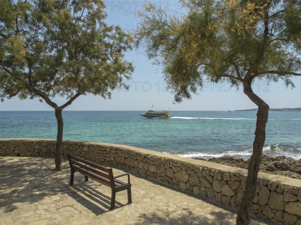 Two trees and a bench on a stone wall by the sea with a passing boat, sa coma, mallorca, spain