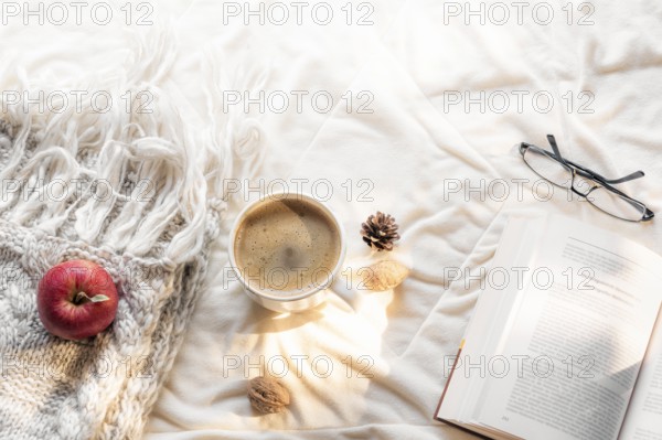 Cozy leisure concept with an open book, glasses, an apple on a fluffy shawl and a hot cup of coffee, on a white blanket, in the morning sun