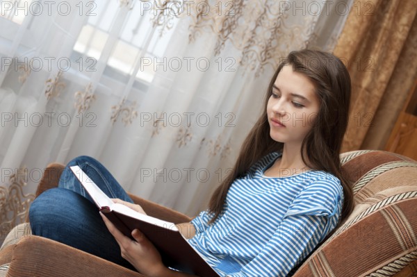 A young caucasian girl reads a book sitting in an armchair