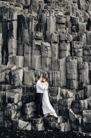 A wedding couple stands on a wall of stone pillars. The bride and groom are hugging on basalt Kekurs, on the black sandy beach of Vik. Destination Iceland wedding