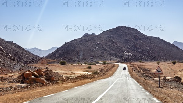 A car drives on a long road through a mountainous desert landscape under a blue sky, By car through the Sahara in Algeria