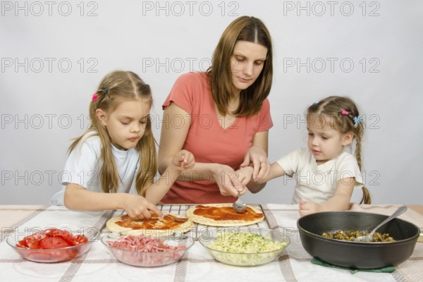 Mom helps younger daughter spread ketchup on a pizza, the eldest daughter, she is preparing a second pizza