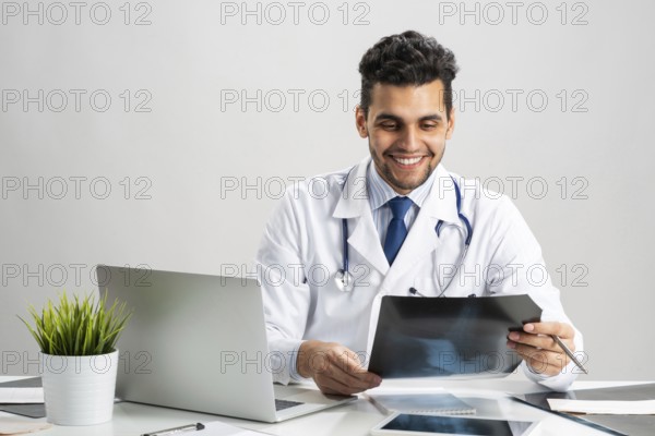 Handsome doctor looking at x-ray scan. Happy physician in white coat with stethoscope sitting at desk with laptop. Professional medical diagnosis and treatment in hospital. Medical healthcare services