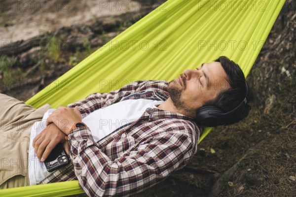 Man on bicycle trip at camping by lake is relaxing in green hammock while listening to music. Active recreation theme in nature. Hipster cyclist with headphones having fun in hammock by river