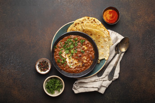 Traditional Indian Punjabi dish Dal makhani with lentils and beans in black bowl served with naan flat bread, fresh cilantro and two spoons on brown concrete rustic table top view. Space for text, food photography, food photography