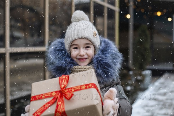 Portrait of joyful girl with a gift box for Christmas on a city street in winter with snow on a festive market with decorations and lights. Warm clothes, knitted hat, scarf and fur. Copy space