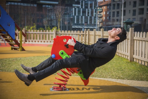 Young man reliving his childhood plying in a childrens playground riding on a colourful red spring seat with a happy smile in an urban park