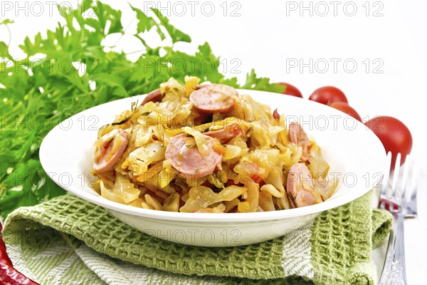 Cabbage stew with sausages in a white plate on a towel, tomatoes, parsley and a fork on the background of wooden boards