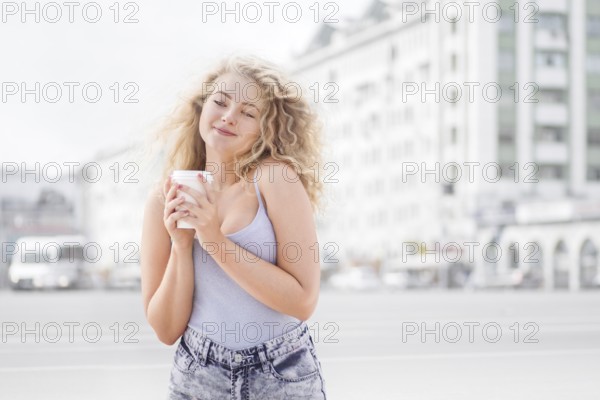 Happy young woman with long curly hair holding a takeaway coffee cup, smiling and flirting with the camera against the backdrop of city traffic