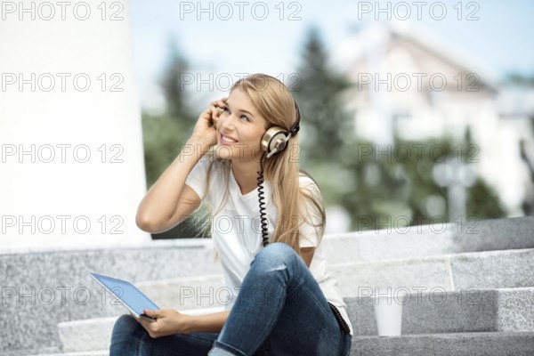 Happy young woman with vintage headphones and a cup of coffee to go, surfing the internet on a tablet PC, listening to music and sitting on a staircase in front of an urban background