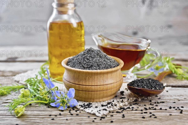 Flour of black caraway in a bowl, seeds in a spoon, oil in a bottle and sauceboat on burlap, sprigs of Nigella sativa with blue flowers and leaves on wooden board