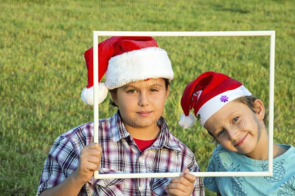 Two brothers in joke caps Santa Claus have fun smiling at the audience, holding a white frame. Background is a green summer park. Concept - portrait and promotional photo