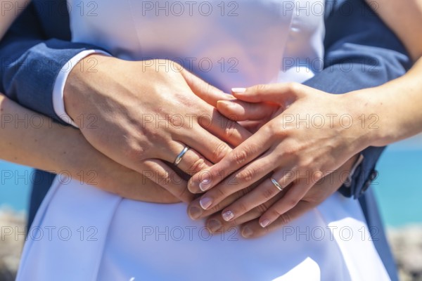 A couple is holding hands and wearing wedding rings. The woman is wearing a white dress and the man is wearing a blue suit. Scene is romantic and happy