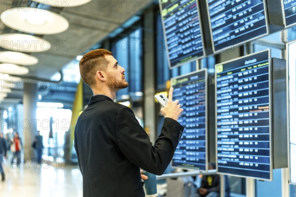 Businessman holding smartphone and pointing at timetable display, checking flight information in airport terminal
