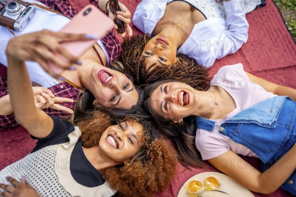 Four cheerful female friends are lying in a circle on a red blanket outdoors, taking a selfie with a smartphone