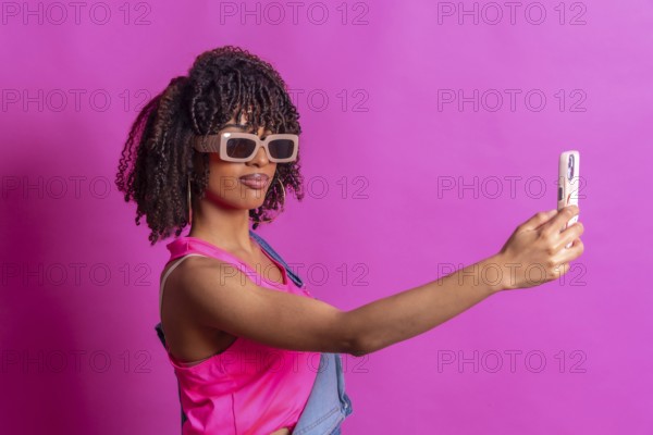 Fashionable young woman taking a selfie with smartphone, wearing sunglasses and pink top on a vibrant pink background
