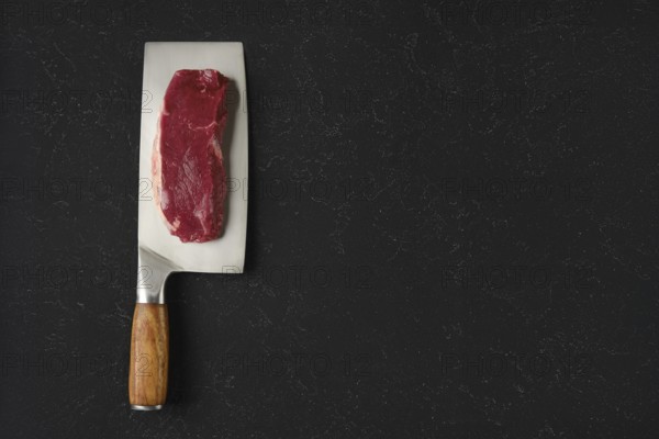 A piece of raw beef meat on a sharp cleaver with a wooden handle. The cleaver rests on a black countertop, showcasing the texture of the meat and the gleam of the blade
