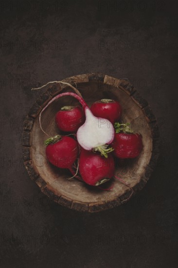 A collection of vibrant red radishes sits in a rustic wooden bowl. The contrasting dark background highlights the freshness and natural beauty of the vegetables, showcasing their green tops.