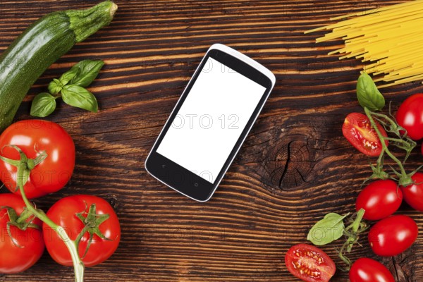 Fresh vegetables and pasta on wooden table with smartphone and recipe on screen. Groceries, recipe and cooking. Modern food preparation
