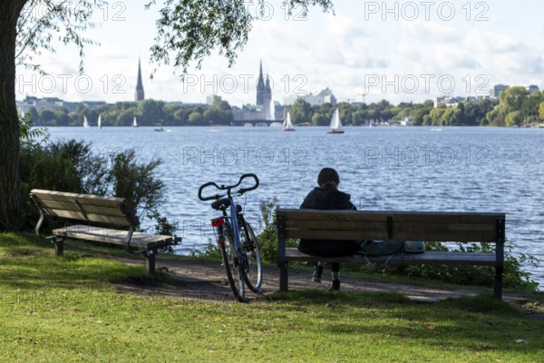 Leisure activities in summer, bicycle and woman on park bench, behind them sailing boats in front of blue sky on the Alster, Hamburg, Germany