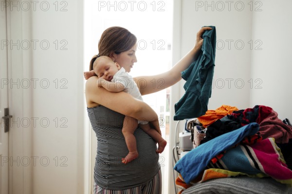 Young mother holding sleepeing baby on her shoulder and trying to sort the laundry simultaniously. Mother combines everyday routines with raising her newborn baby