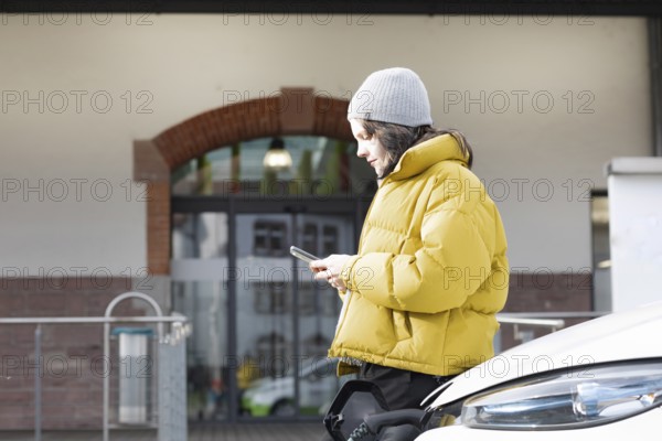 Woman with mobile phone in yellow jacket at charging station with electric car, environmentally friendly