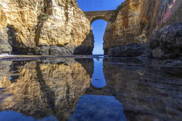 Yellow rocks and cliffs on a sandy beach. A bay by the sea, over which the old stone bridge of the Pinhão fortress connects two cliffs. Praia da Caldeira, Lagos. Algarve, Portugal