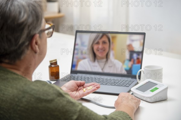 Senior woman having online medical consultation with doctor using laptop, holding pills in hand and checking blood pressure at home