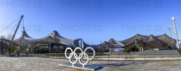 Panoramic photo of the sculpture of the Olympic rings and in the background the Olympic site with the Olympic buildings with blue sky in the background, Munich, Upper Bavaria, Bavaria, Germany