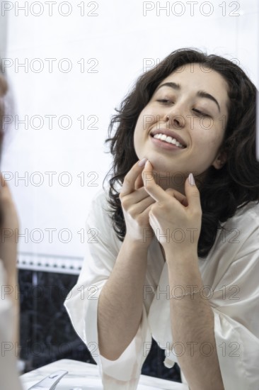 Portrait of a happy woman removing pimples and blackheads from her face in front of the mirror at home. The bad eating routine and the excess of fats and sweets in meals can cause an increase in acne in people