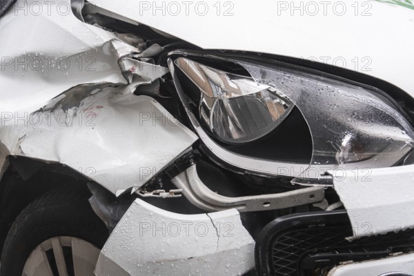 A heavily damaged white car after an accident. The front left mudguard is badly dented and crumpled, while the headlight is partially broken. The body has cracks and deformations, Düsseldorf, Germany