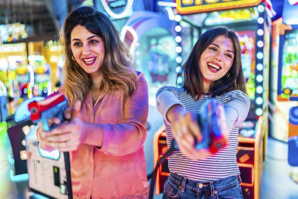 Two young women enjoying their free time playing together in an amusement arcade, holding toy guns and having fun