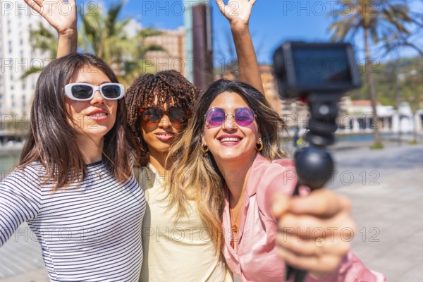 Three cheerful women taking a selfie with action camera, waving and smiling, enjoying a summer day together in the city