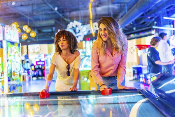Two female friends concentrating while playing air hockey in a vibrant arcade, enjoying their leisure time
