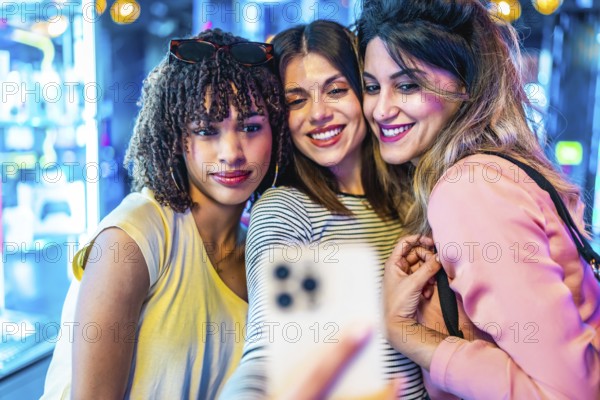 Three cheerful female friends are taking a selfie with a smartphone in a vibrant, illuminated urban setting, possibly an arcade or entertainment venue, enjoying their evening out