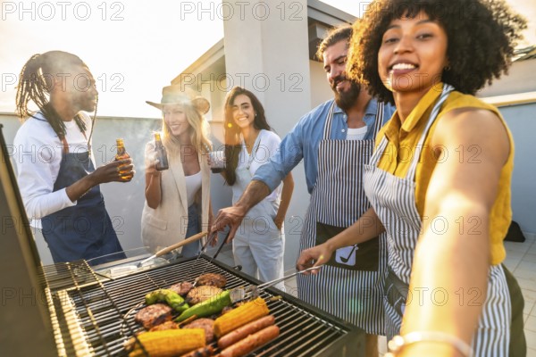 Cheerful multi ethnic friends are having fun, drinking beer and taking selfie while preparing barbecue on a rooftop at sunset