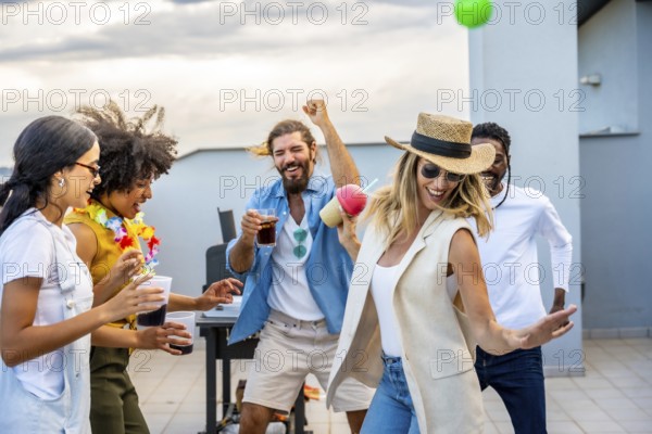 Cheerful multi ethnic friends dancing joyfully and sipping cocktails while celebrating together at an exciting rooftop party during sunset