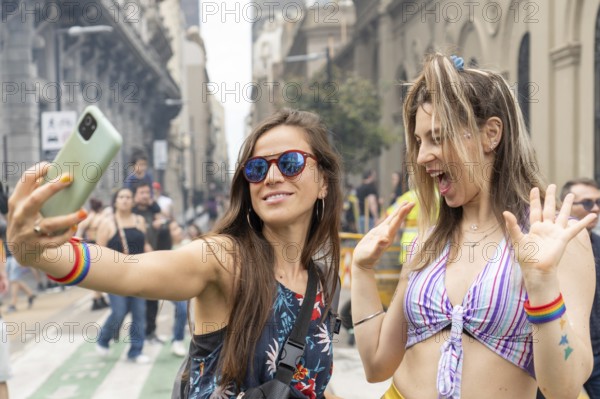 Two women wearing rainbow bracelets are taking a selfie with a smartphone at a gay pride parade, celebrating diversity and inclusion with excitement and joy