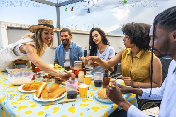 Happy multi ethnic friends having lunch together on urban rooftop, eating hot dogs, burgers, and drinking beer