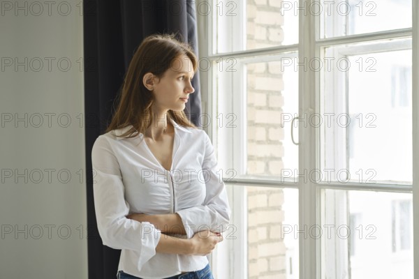 A teenager stands by a window, gazing outside with a thoughtful expression. The cozy room features light curtains and a brick wall, creating a serene atmosphere for reflection and contemplation