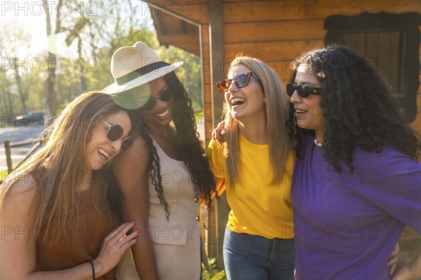 Four diverse women wearing sunglasses and casual clothes are enjoying a sunny day in nature, laughing and embracing each other
