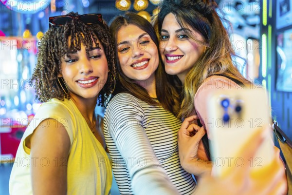 Smiling friends capturing a joyful selfie with a smartphone in a vibrant amusement arcade filled with colorful lights and excitement