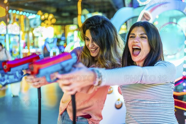 Two women laughing and enjoying their free time while playing arcade games with colorful toy guns, surrounded by vibrant lights and excitement